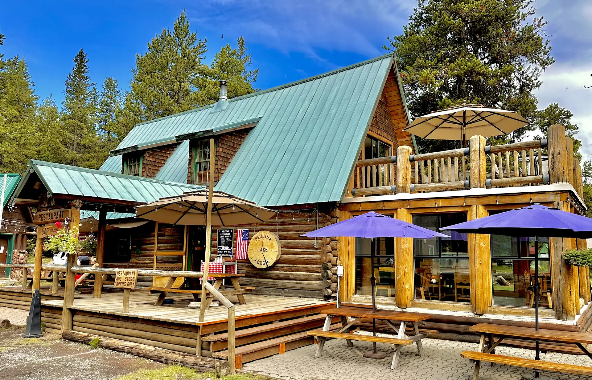 A charming log cabin building with a green roof, outdoor seating, and umbrellas in a scenic forest setting.