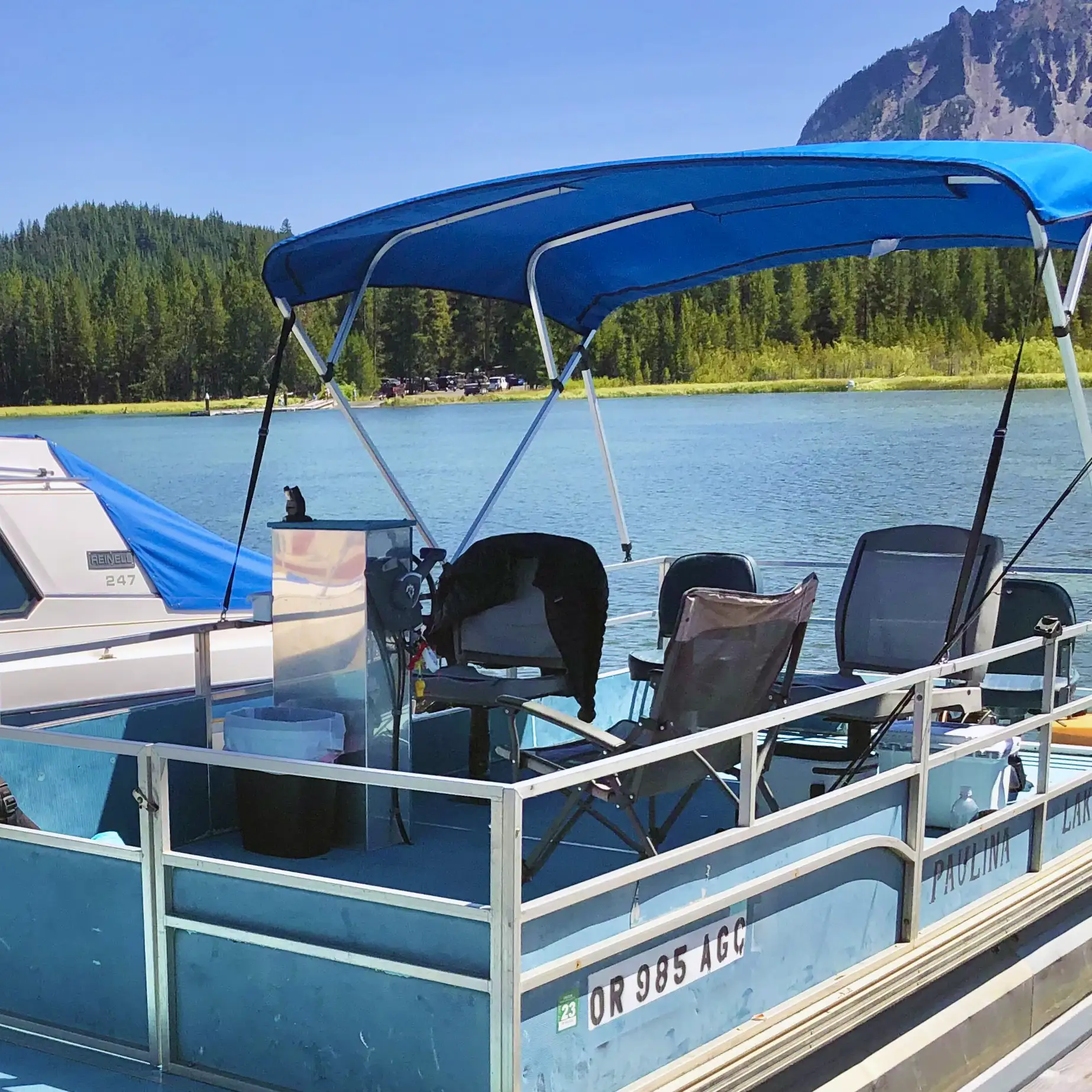 A blue boat with a canopy and two chairs is docked by a tranquil lake surrounded by trees.