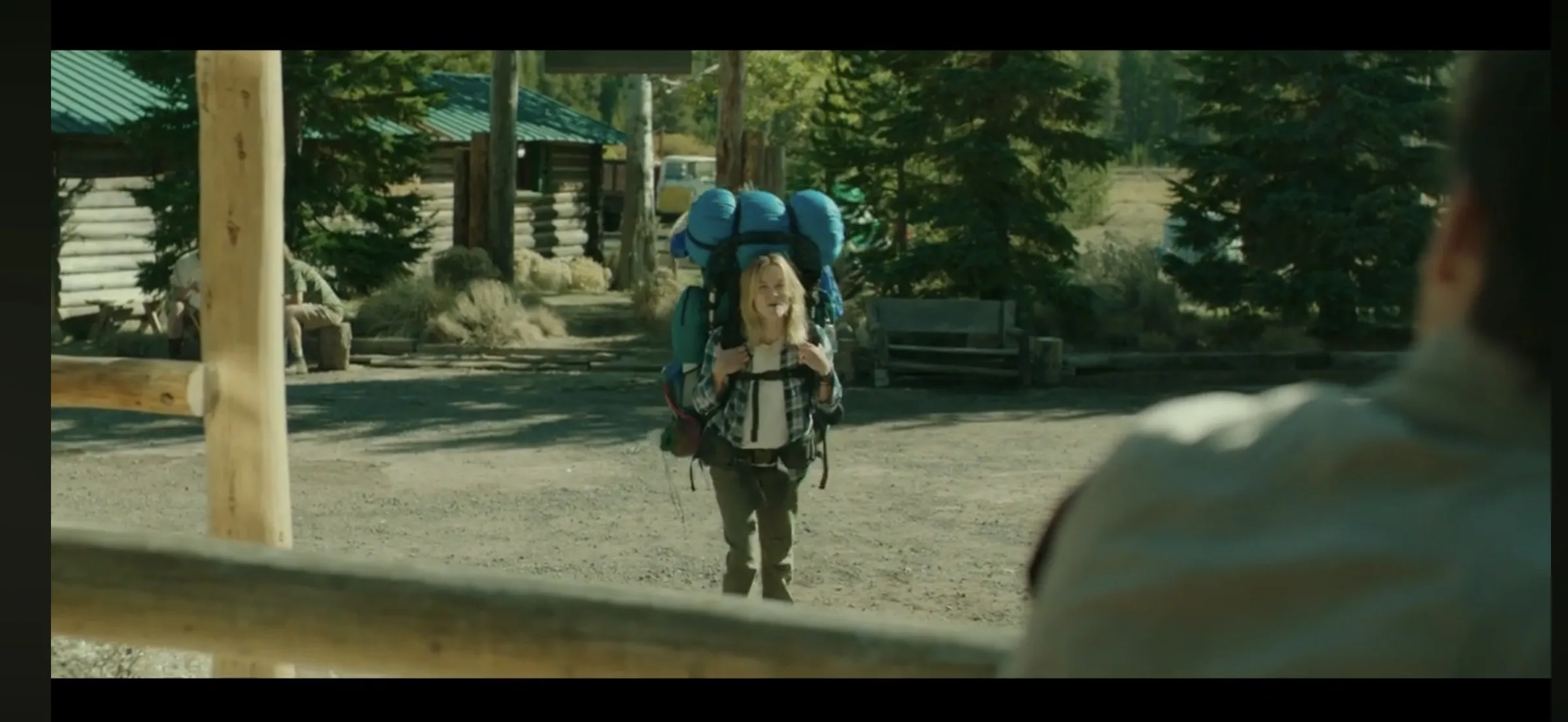 A child with a large backpack stands in front of a rustic cabin in a forested area.