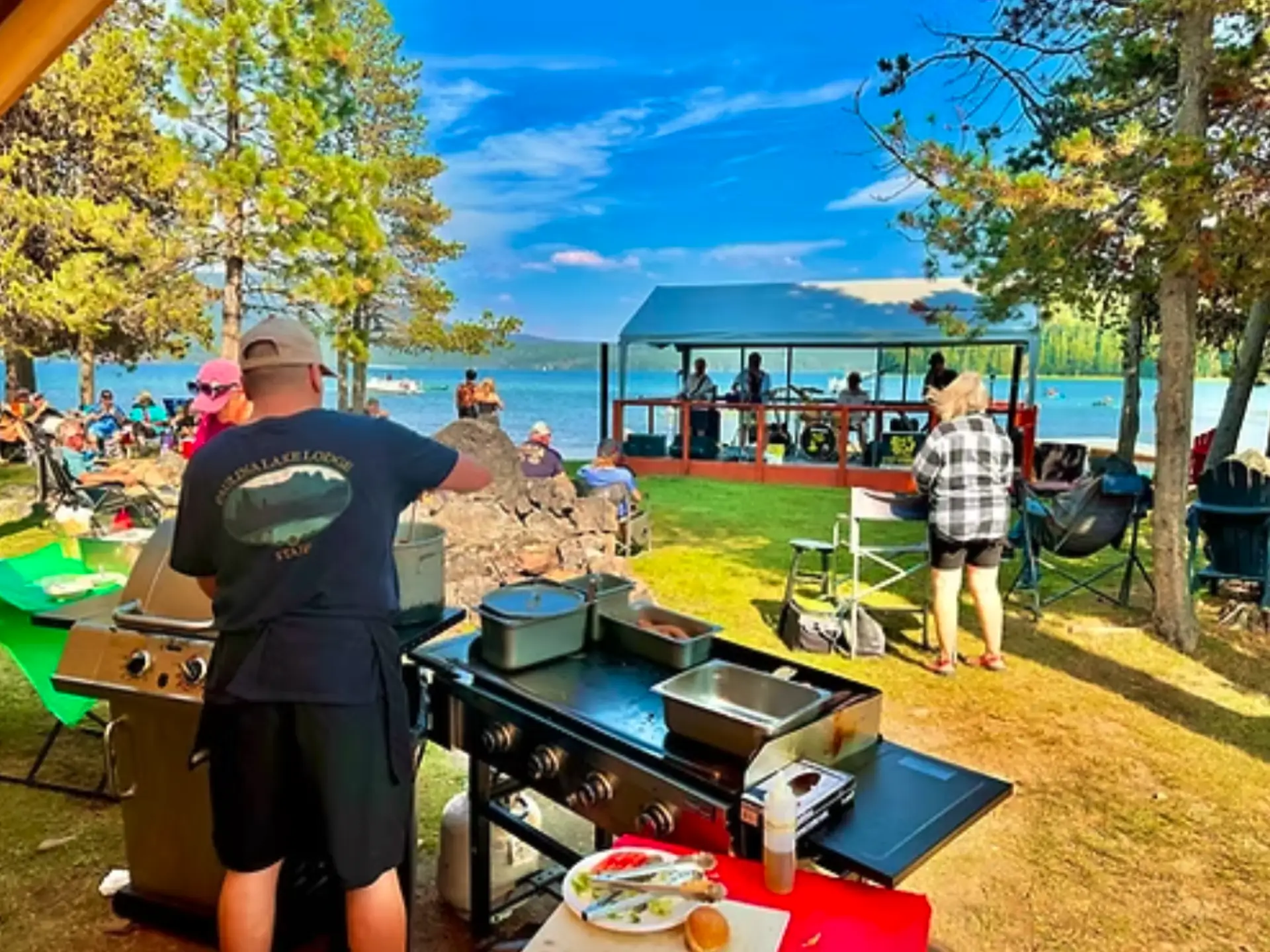 A person cooks on a grill near a lake, with people gathered in the background enjoying the outdoor setting.