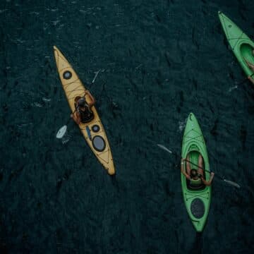 Three kayakers paddle in dark water, two in green kayaks and one in orange.