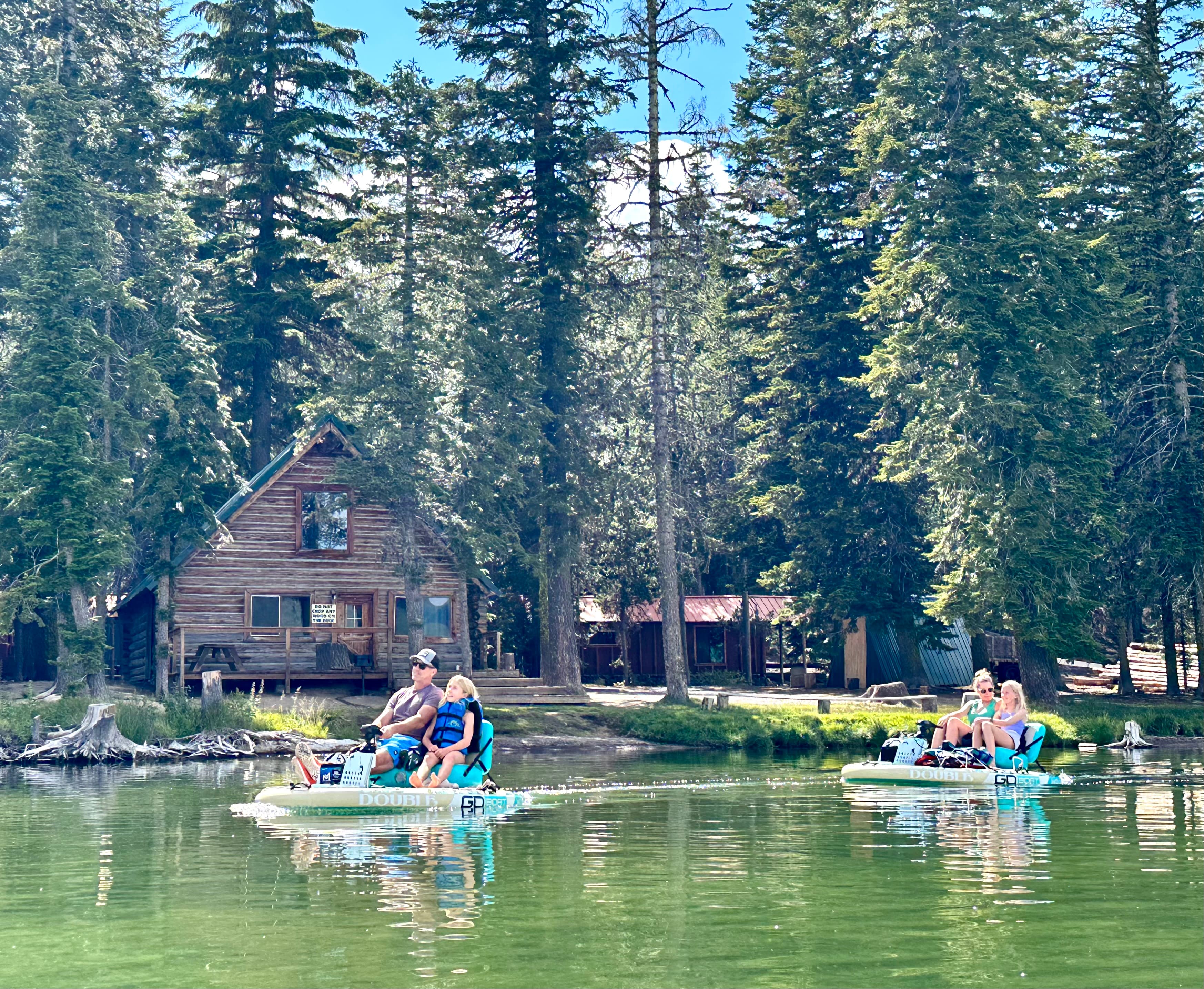 Two pairs of people paddle in small boats on a lake surrounded by trees and a rustic cabin.