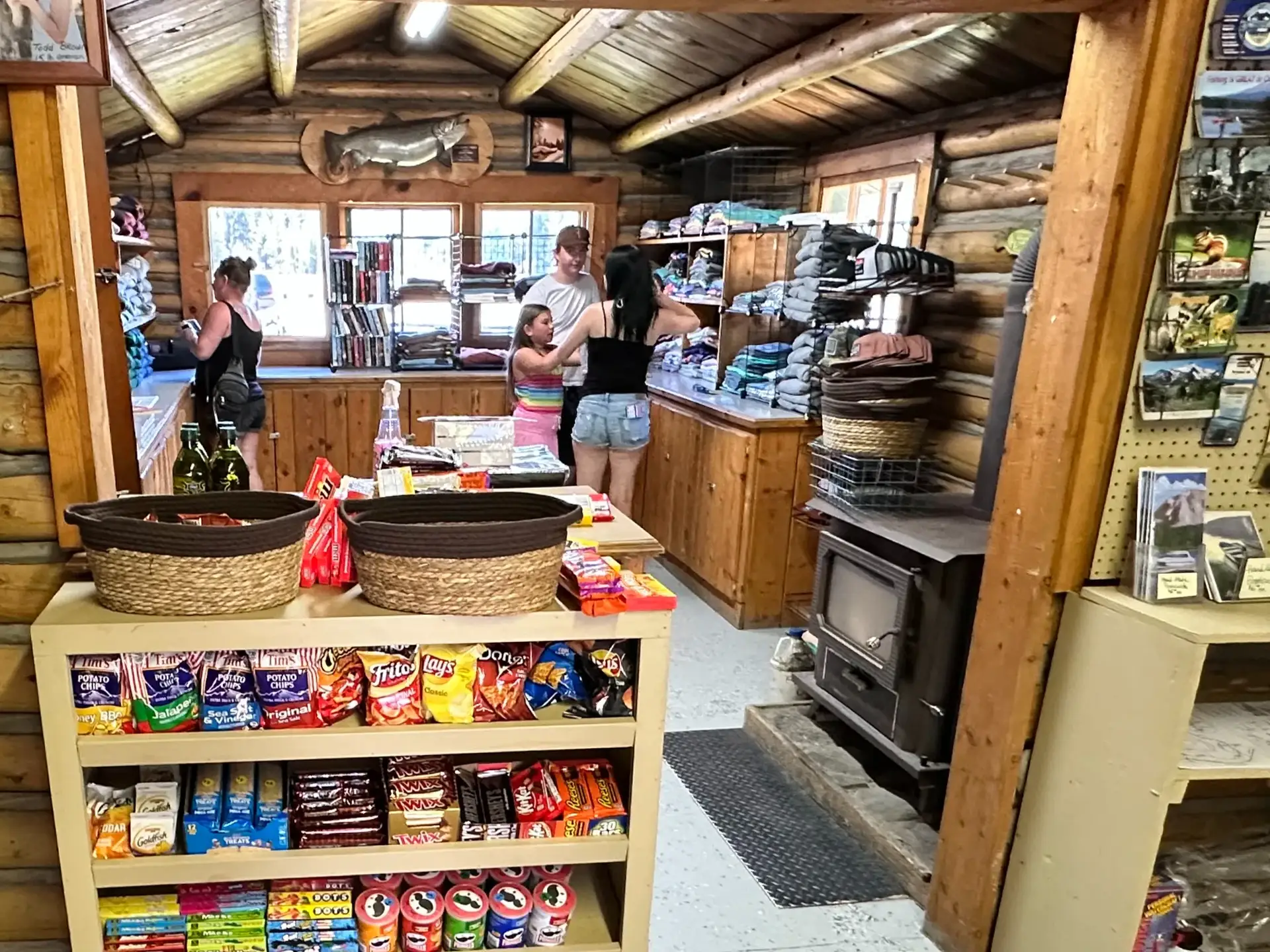 Interior of a rustic shop displaying snacks and merchandise, with customers browsing and a wood stove in the corner.
