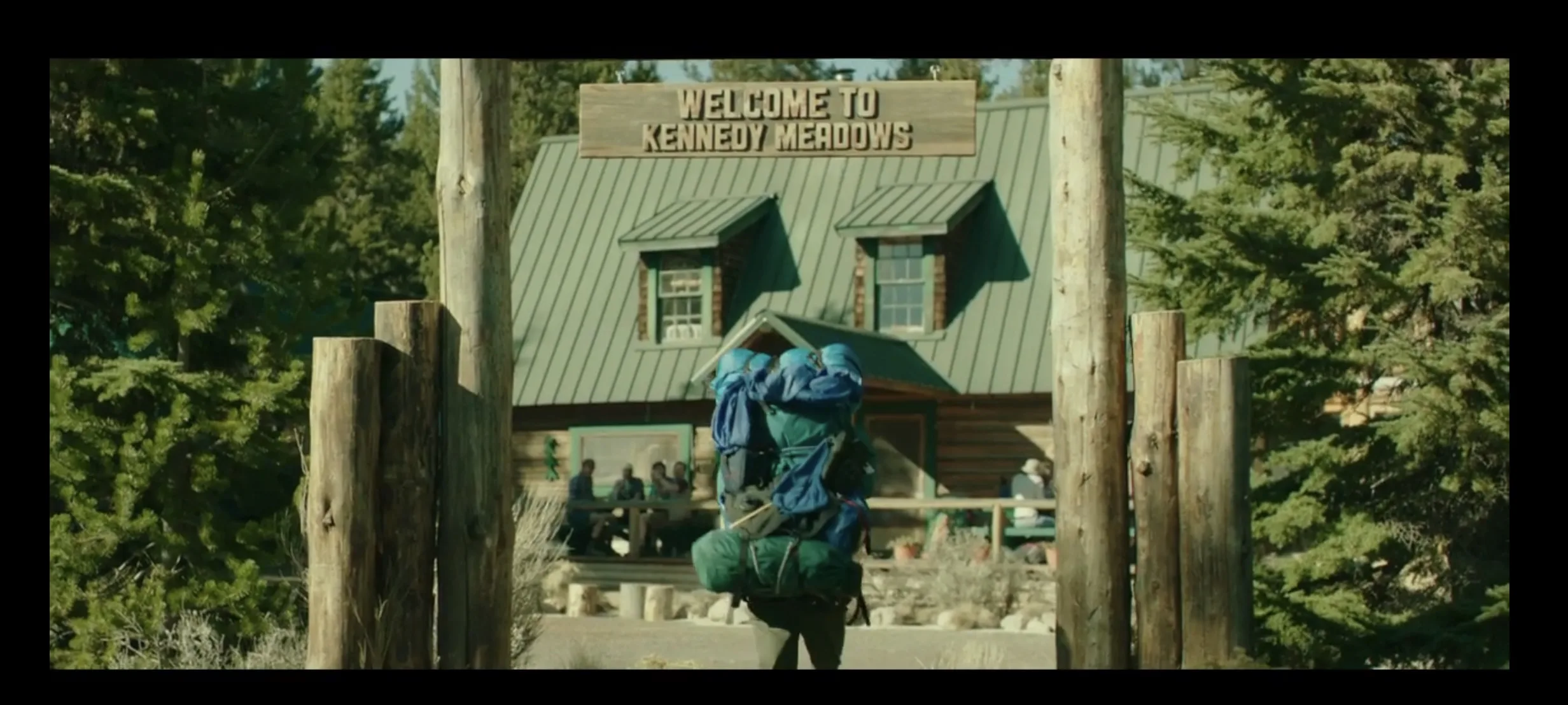 A person carrying a large backpack walks towards a sign that reads "Welcome to Kennedy Meadows," with a cabin and trees in the background.