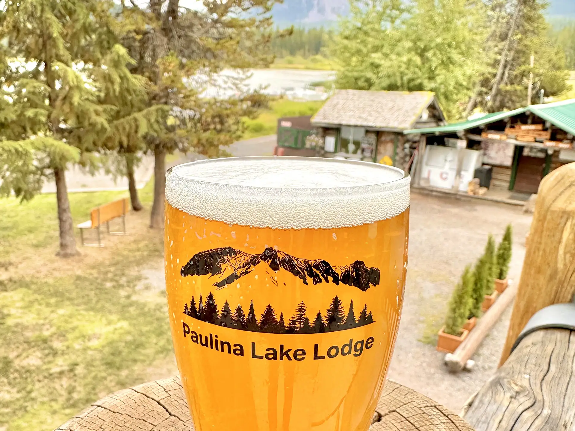 A pint of beer with the "Paulina Lake Lodge" logo sits on a wooden railing, surrounded by trees and a rustic lodge in the background.