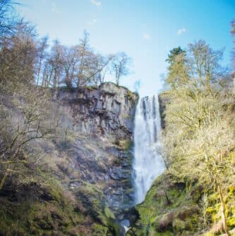 A tall waterfall cascades down a rocky cliff surrounded by lush trees under a blue sky.