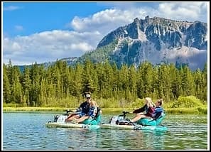 Three people on pedal boats enjoying a lake surrounded by trees and mountains.