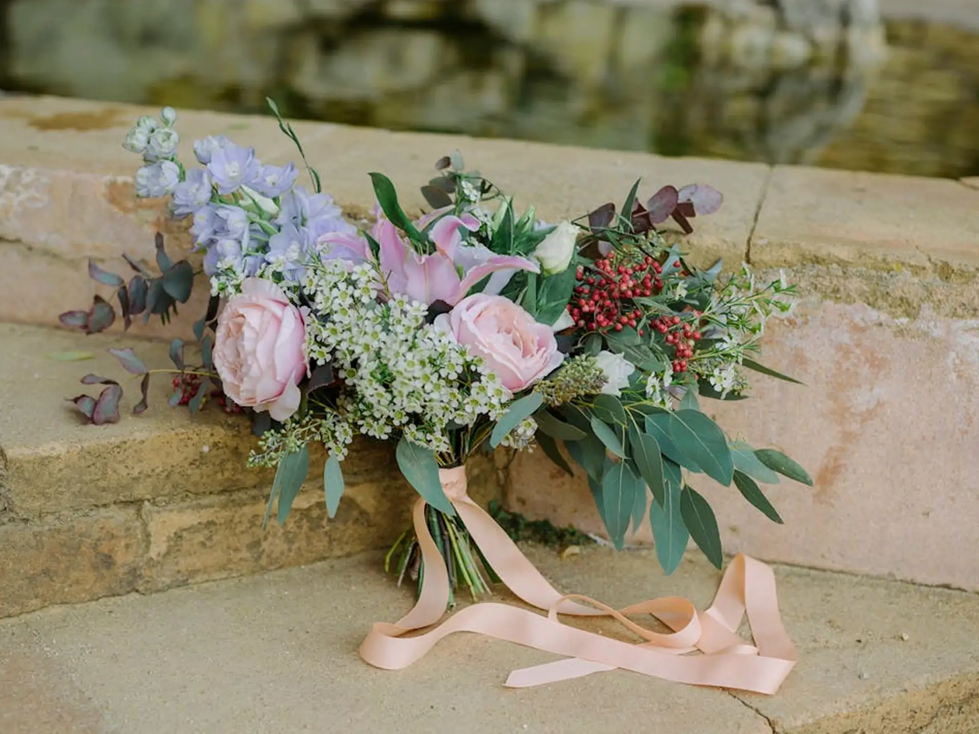 A colorful bouquet of pink roses, lavender flowers, and greenery tied with a ribbon rests on stone steps.