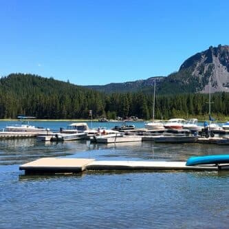 A peaceful marina with boats docked in calm water and a mountainous backdrop under a clear blue sky.