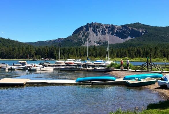 A serene lakeside scene with boats docked and a mountainous backdrop under a clear blue sky.
