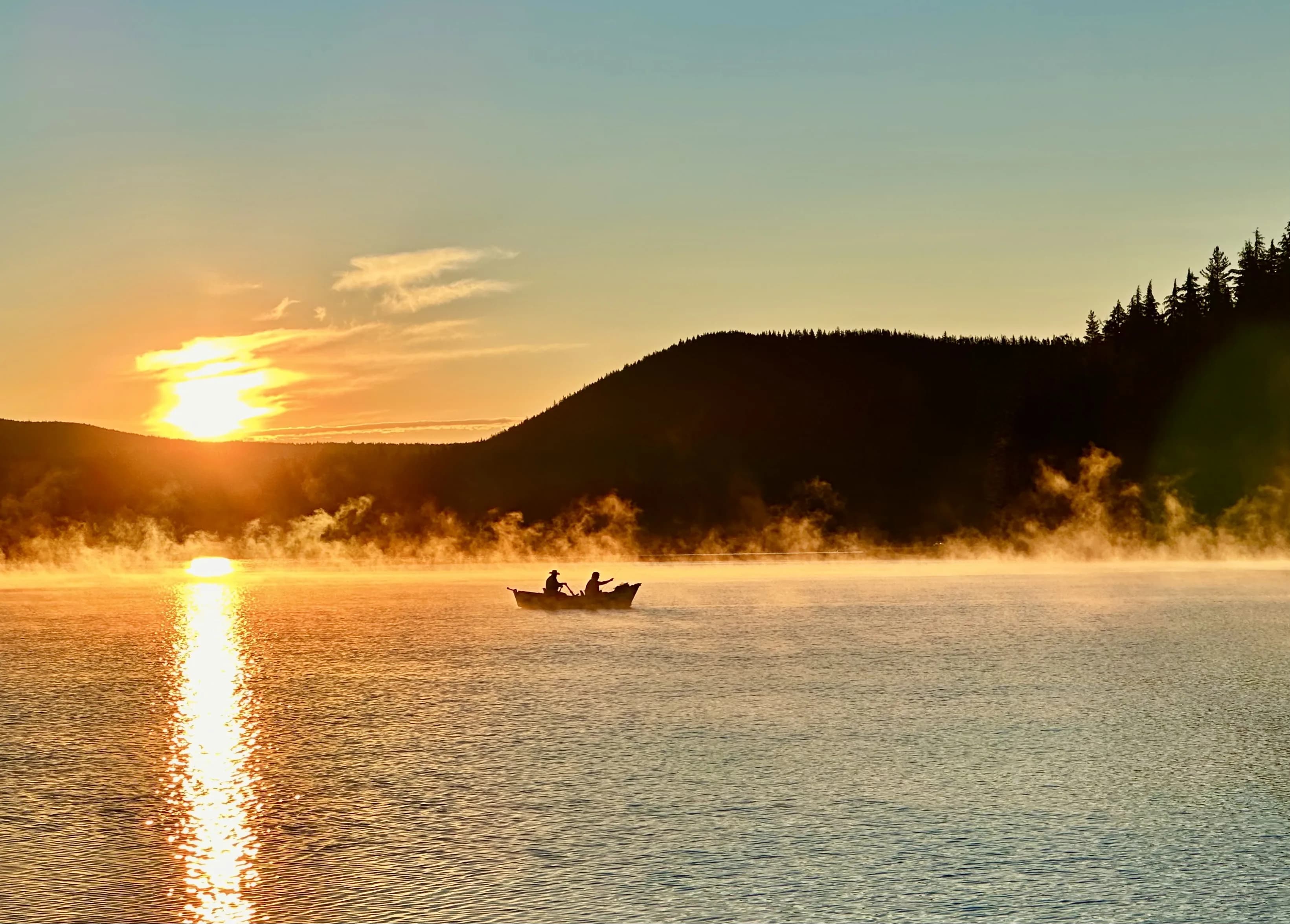 A small boat with two people floats on a misty lake at sunrise, surrounded by silhouette mountains.