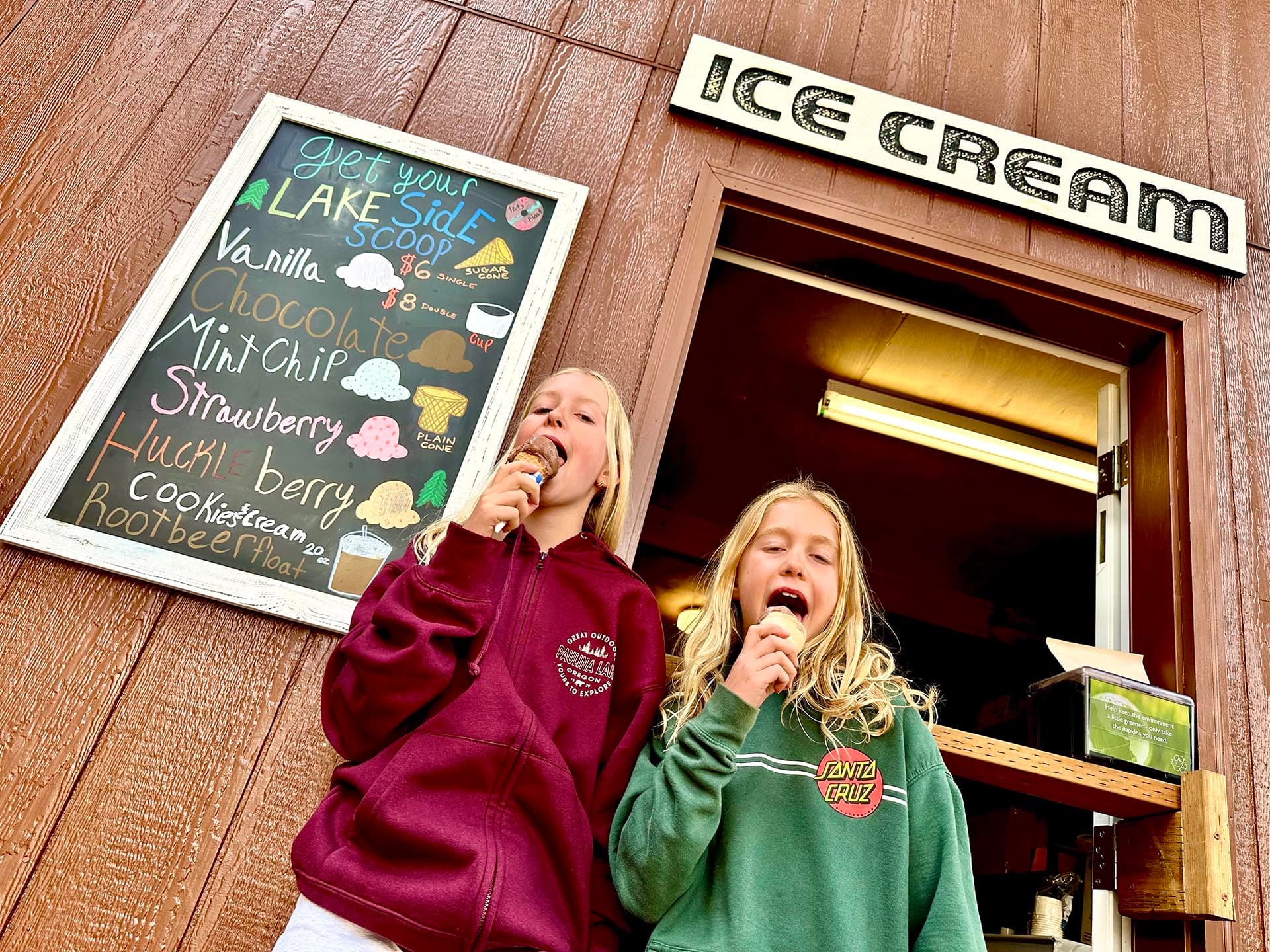Two girls enjoying ice cream in front of an ice cream shop's menu board.