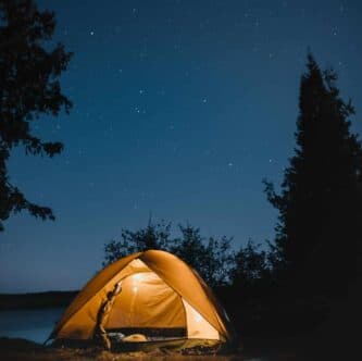 A yellow tent illuminated at night under a starry sky near a body of water.