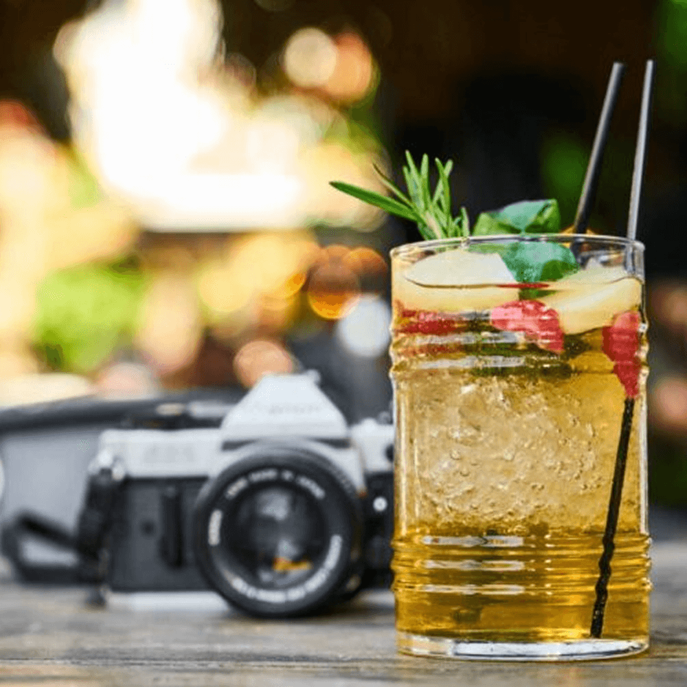 A glass of refreshing drink garnished with fruit and herbs, set on a table beside a vintage camera.