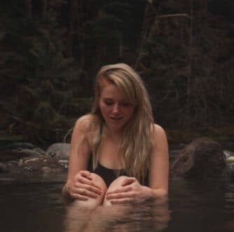 A woman with long blonde hair sits in a serene, rocky stream, looking contemplatively at the water.