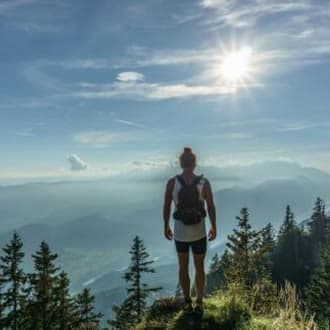 A hiker stands on a mountaintop, gazing at a vast landscape under a bright sun.