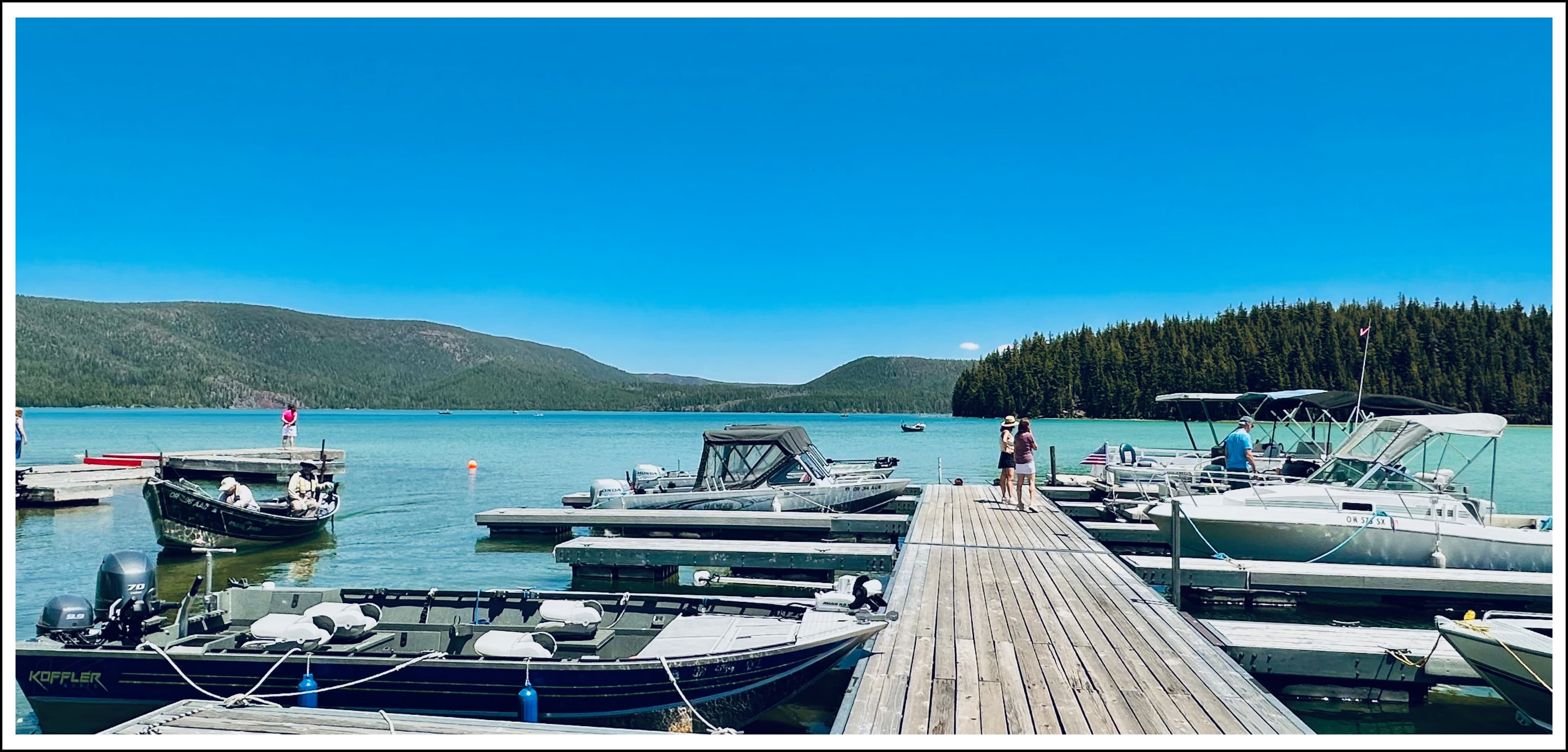 A wooden dock lined with boats near a tranquil blue lake and forested hills under a clear sky.