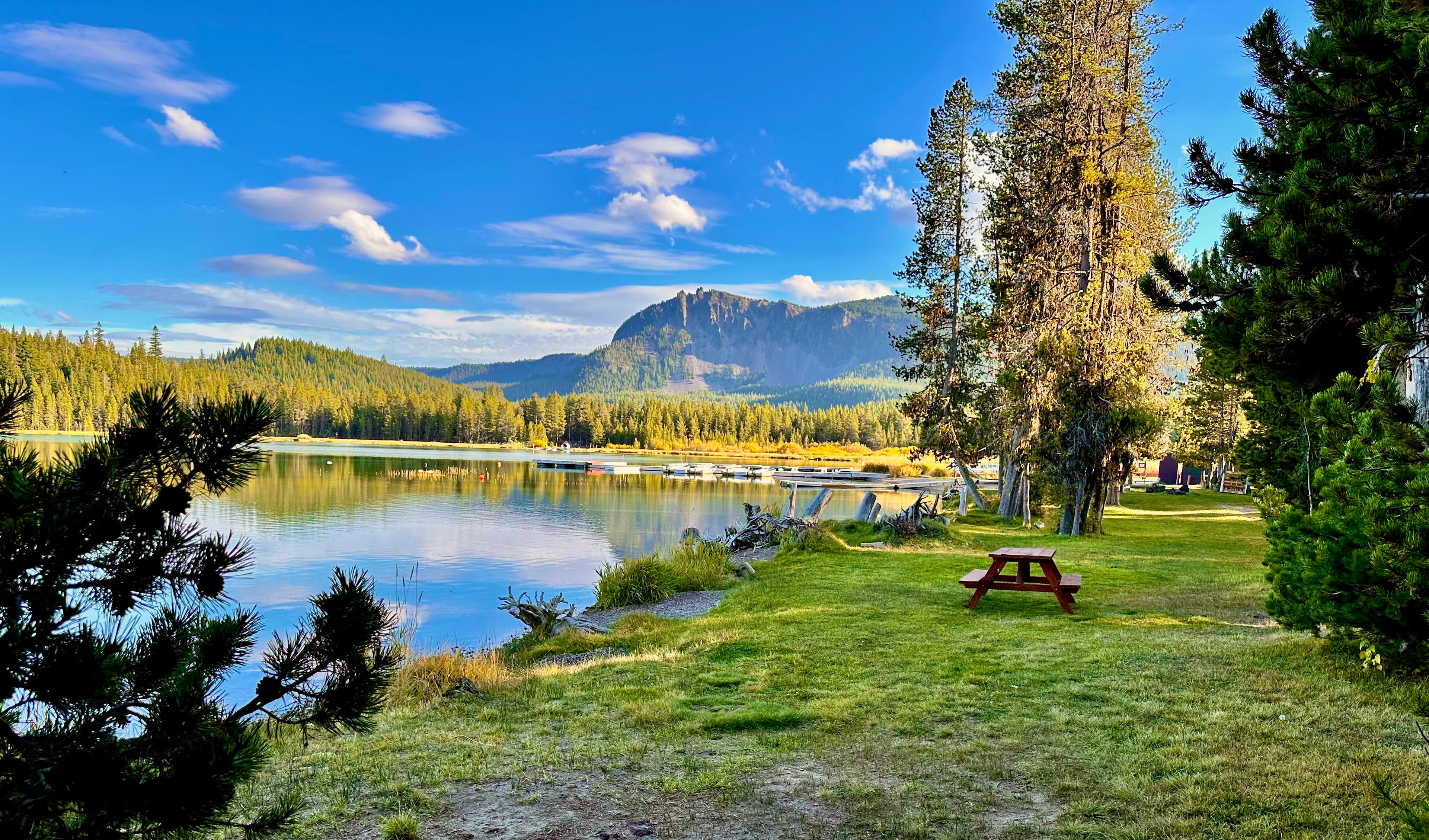 A tranquil lakeside scene with picnic table, surrounded by trees and mountains under a blue sky.