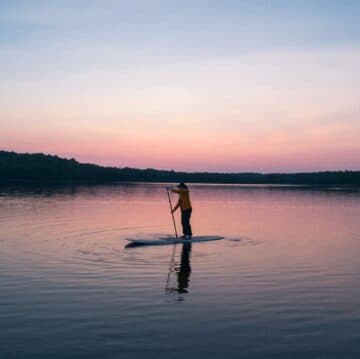 A person paddleboards on a calm lake during a pastel sunset.