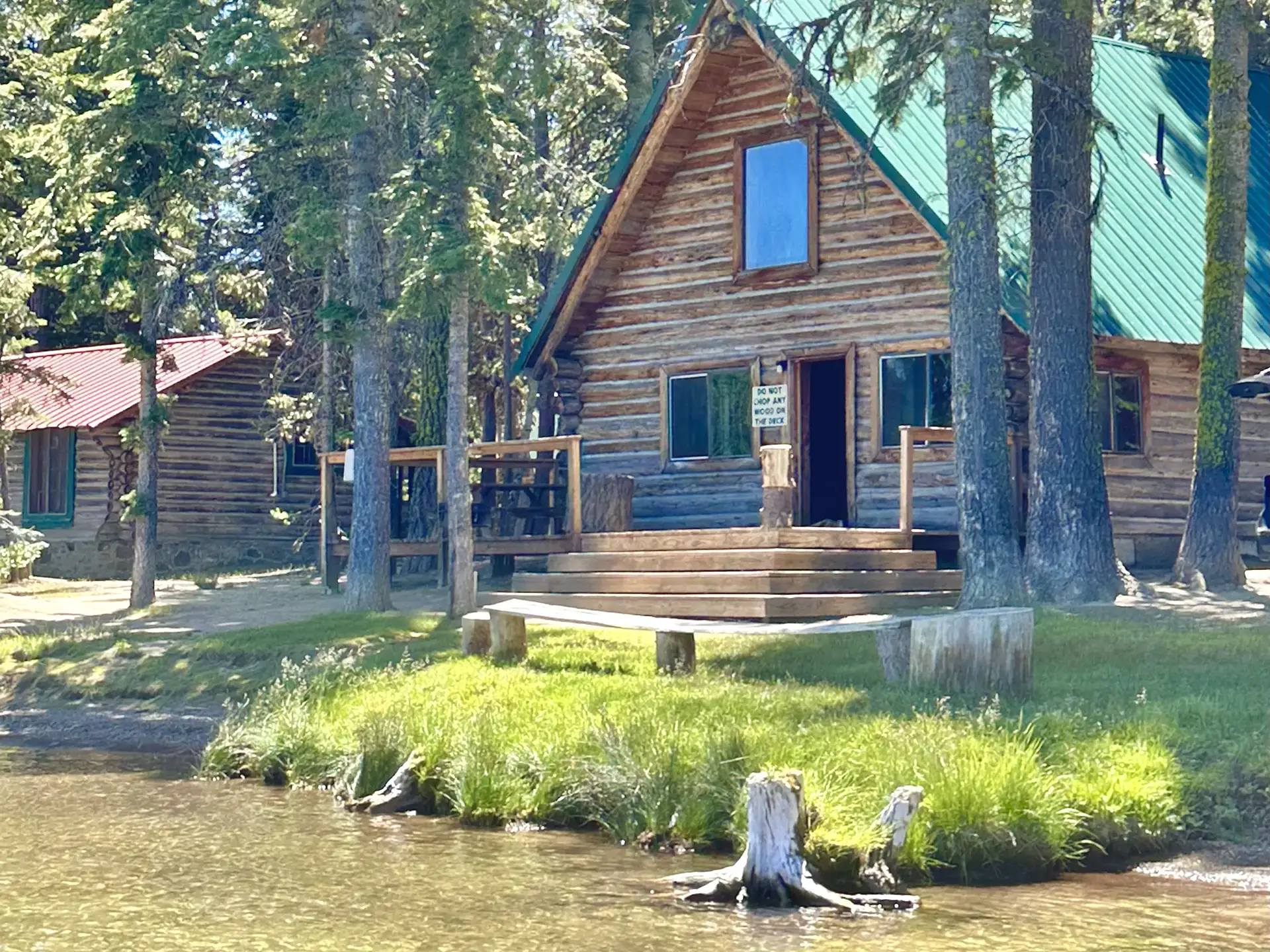 A rustic log cabin with a green metal roof sits by a lake, surrounded by trees.