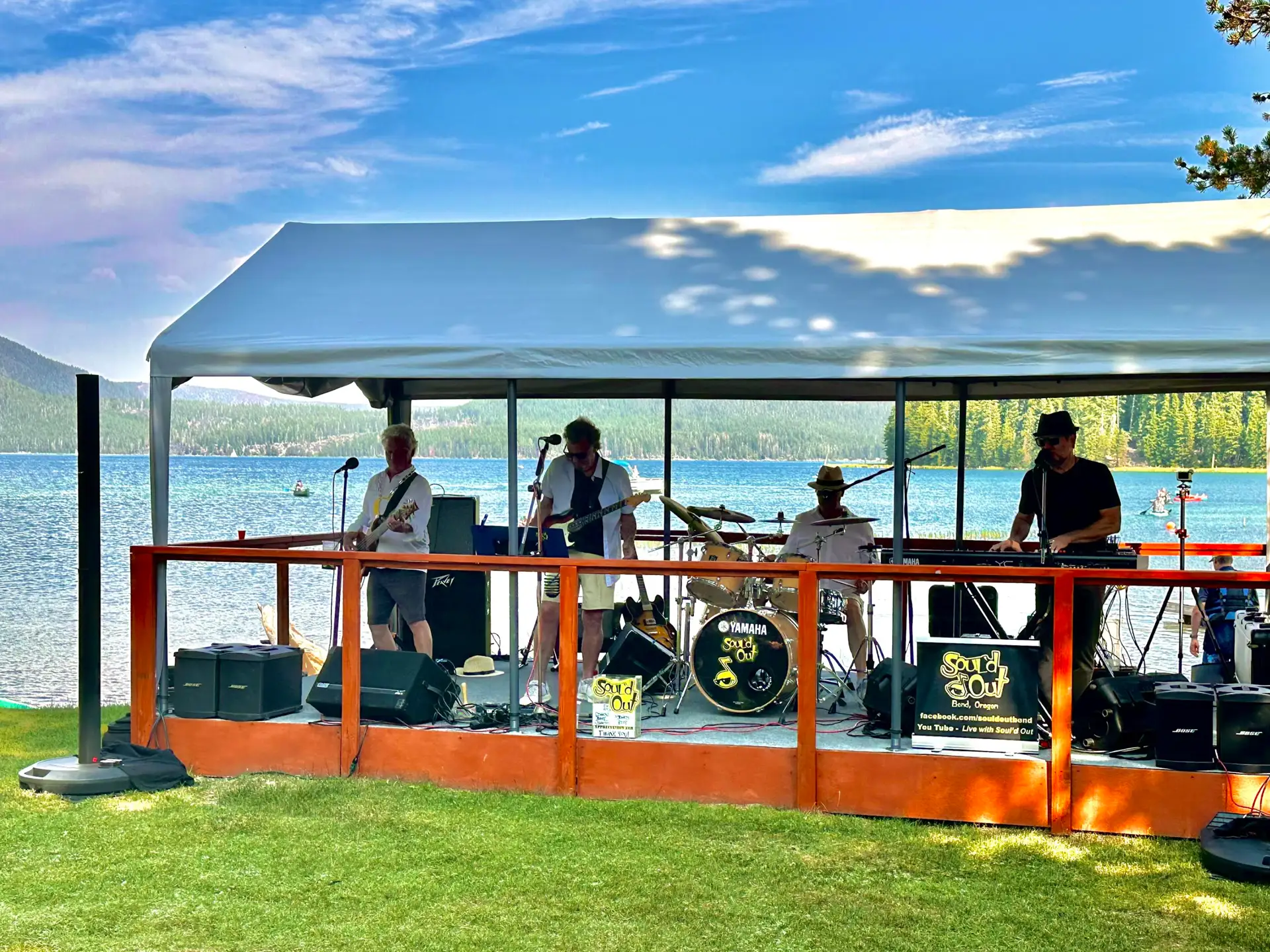 A band performs on a stage by a lake under a tent.
