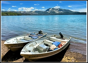 Two boats anchored on a sandy lake shore with a mountainous background.