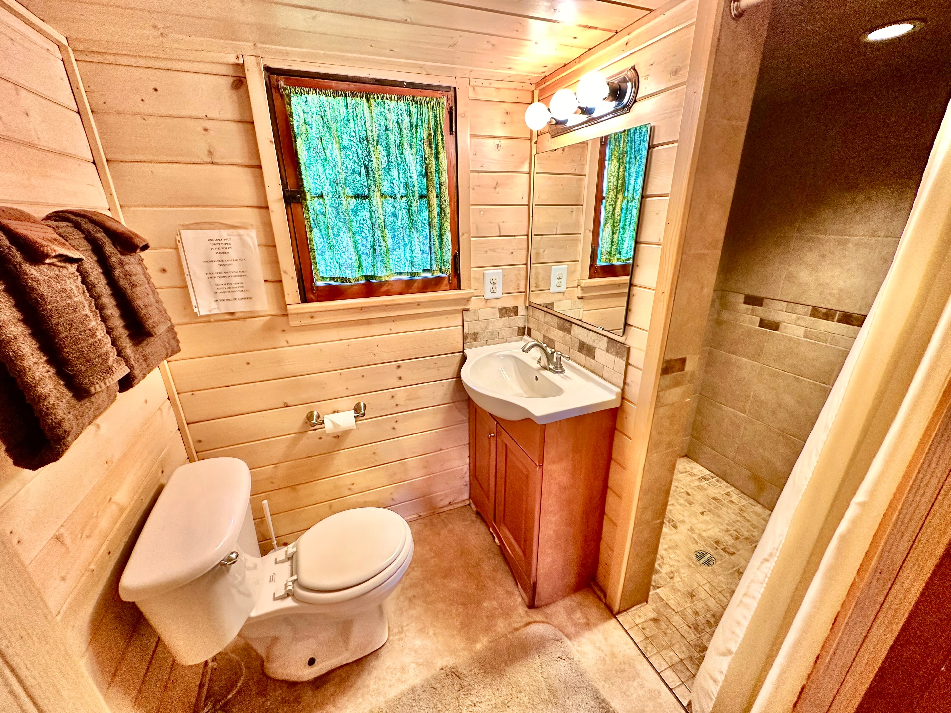 A cozy wooden bathroom featuring a toilet, sink, and shower with natural light streaming through a window.