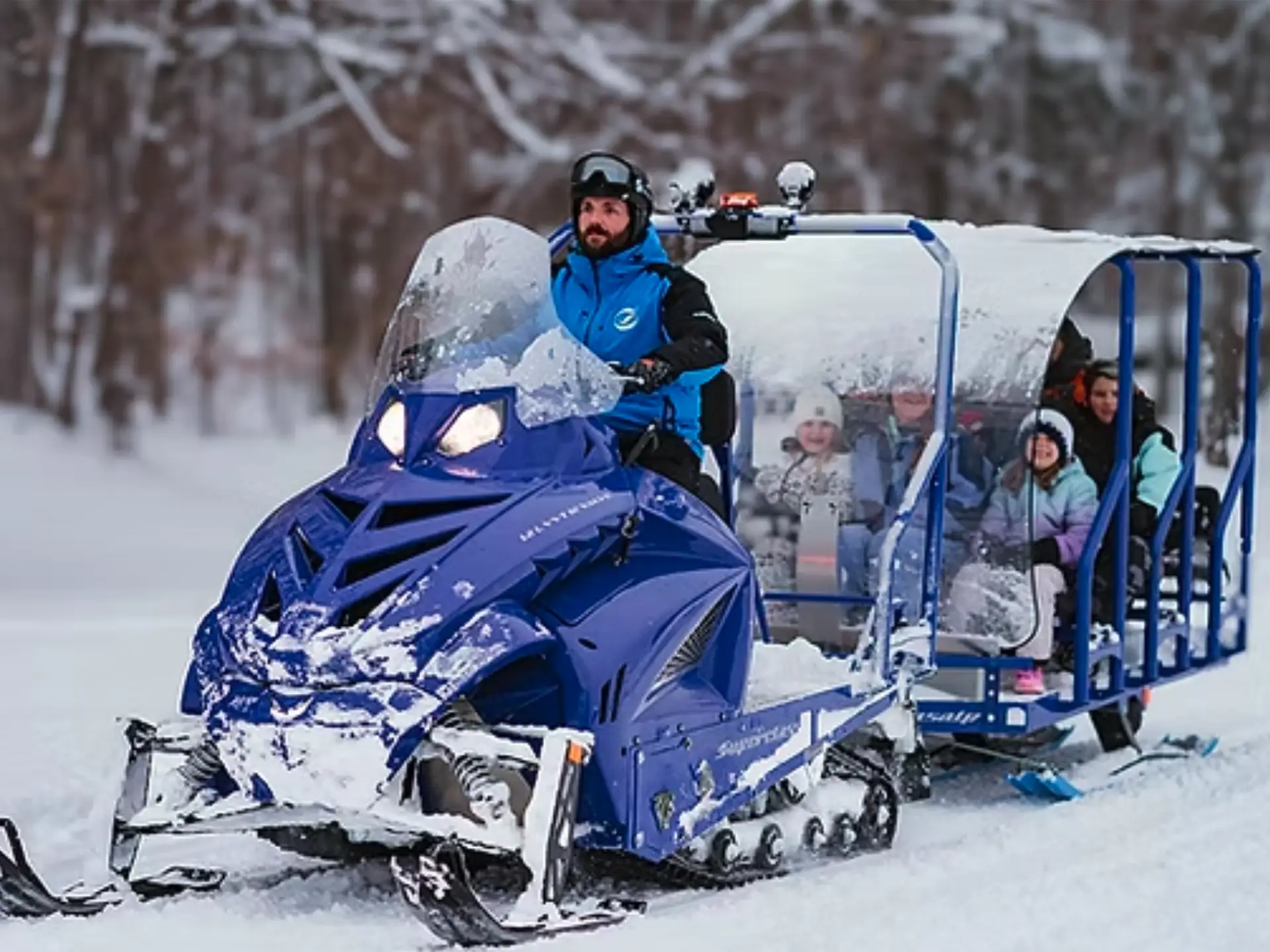A man drives a blue snowmobile towing a covered sled with passengers through a snowy landscape.