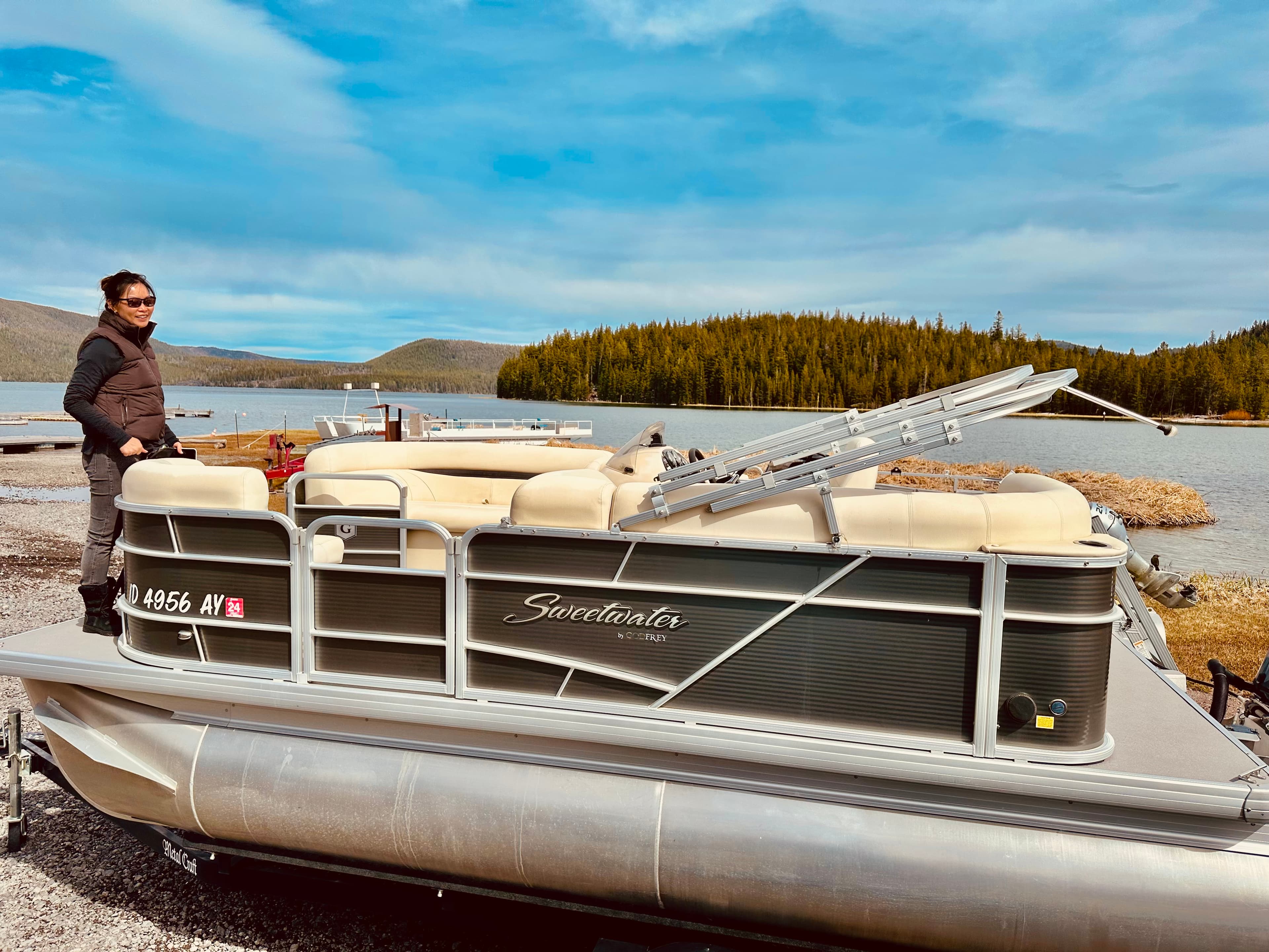 A person stands on a pontoon boat by a lake surrounded by forested hills under a partly cloudy sky.