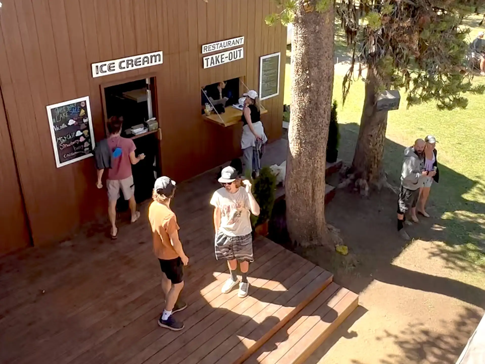 A group of people interacts near an ice cream stand and a take-out restaurant in a sunny outdoor setting.