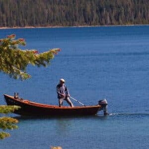 A person is standing in a small boat on a calm lake surrounded by trees.