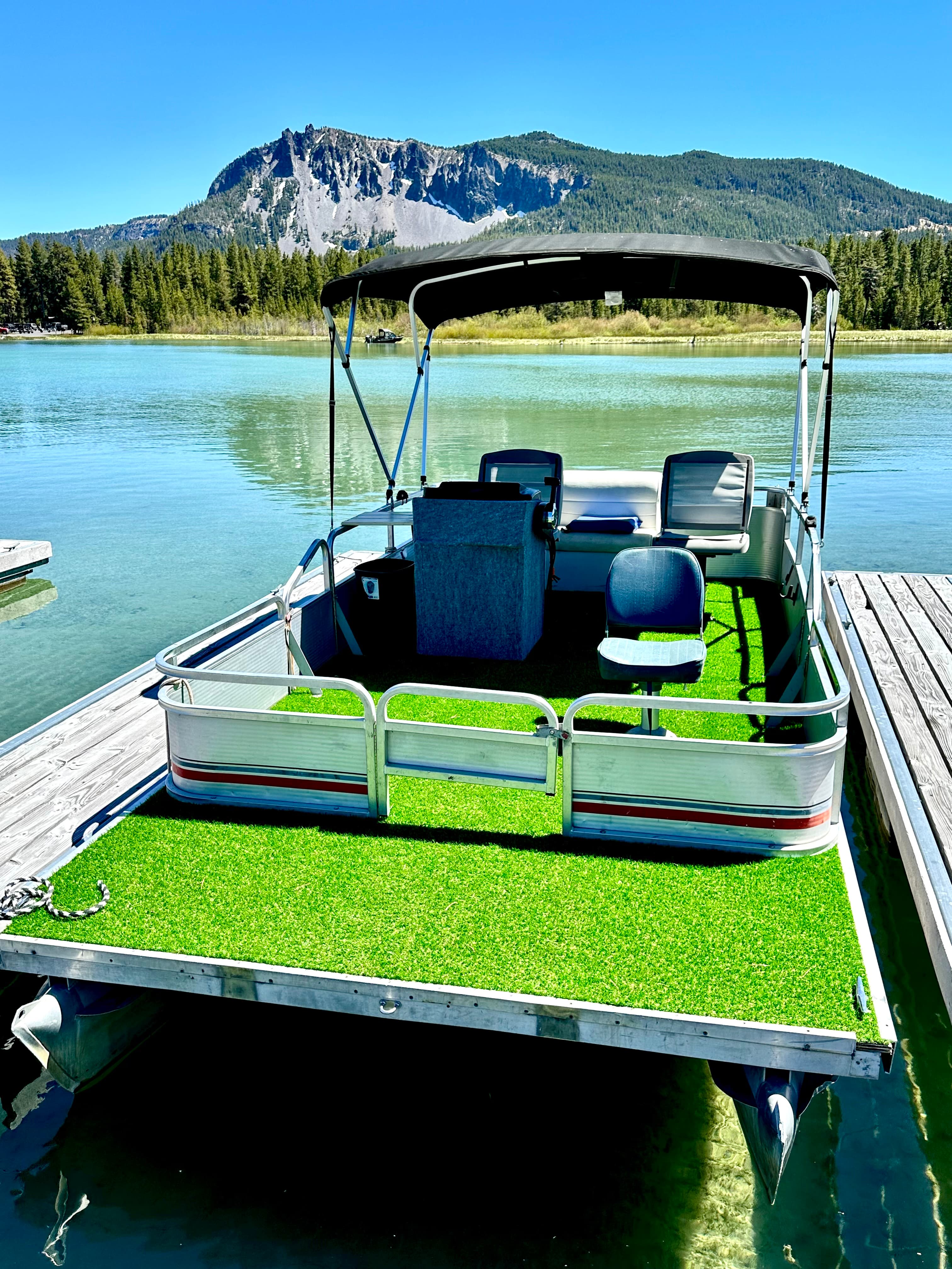 A pontoon boat with artificial turf flooring is docked by a serene lake surrounded by forested mountains.