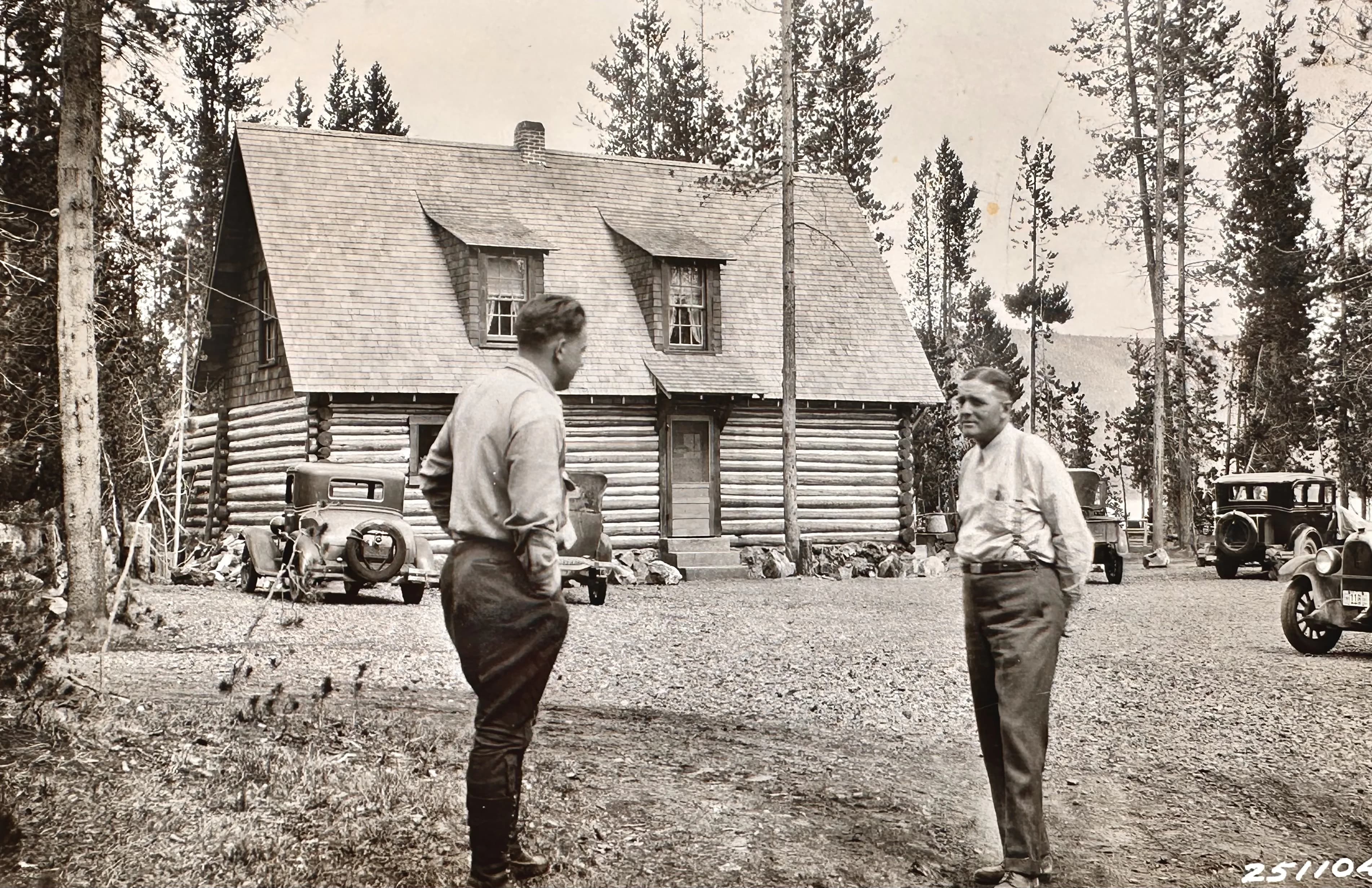 Two men converse outside a log cabin with vintage cars parked nearby.