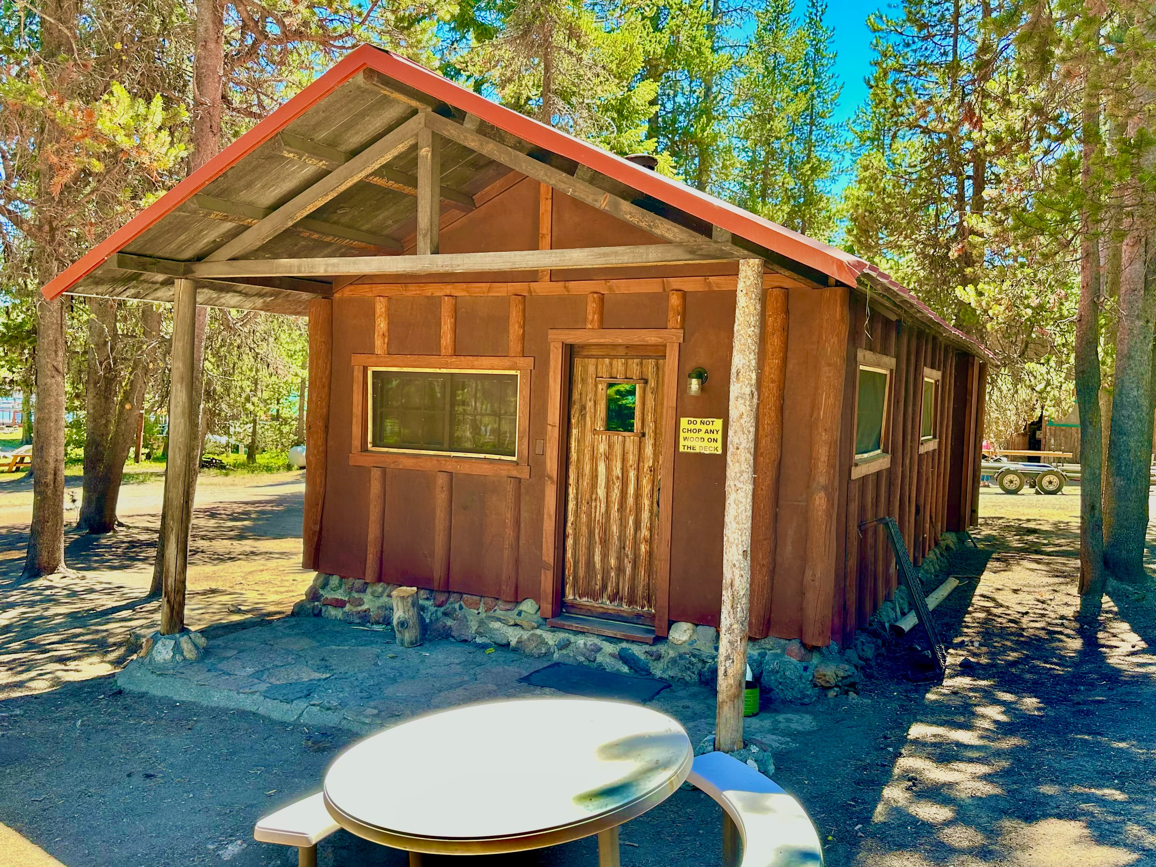 A rustic wooden cabin with a red roof surrounded by trees and a picnic table in front.