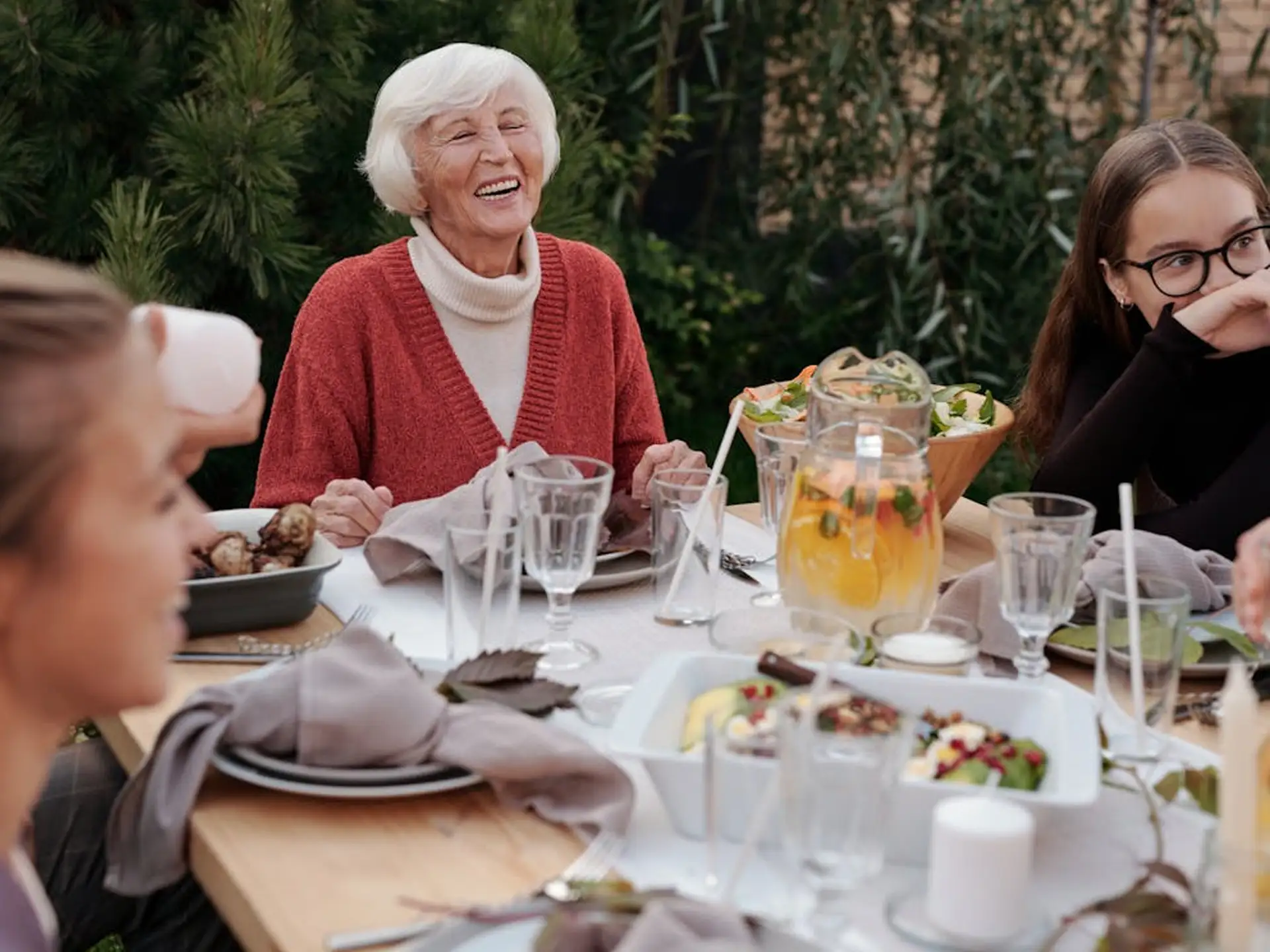 A group of people enjoying a lively outdoor dinner, featuring an elderly woman laughing at the table.